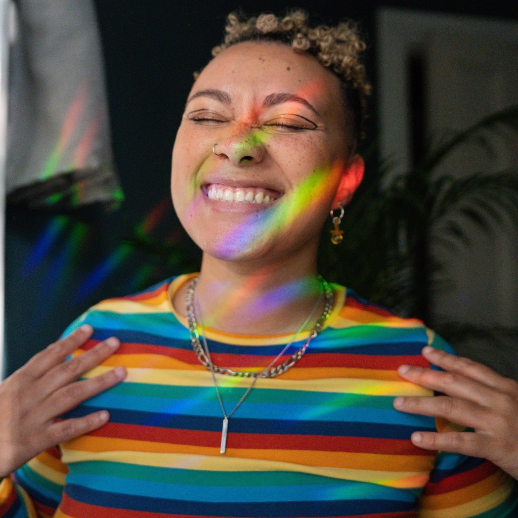 Person with short curly hair wearing a colorful striped shirt, smiling with eyes closed, surrounded by rainbow light reflections.