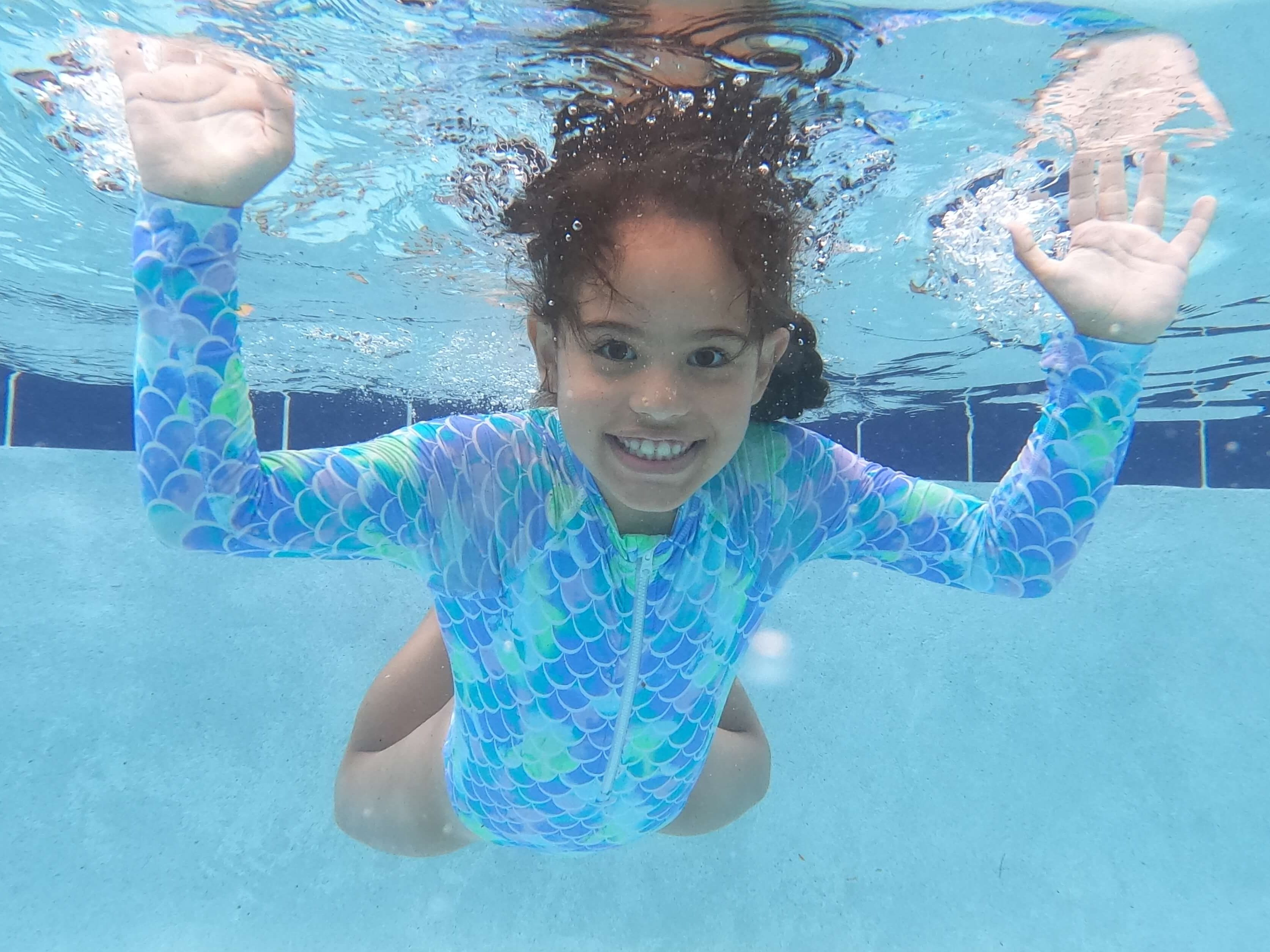 A child underwater smiling and waving, wearing a colorful mermaid-themed swimsuit, with clear blue water surrounding her.